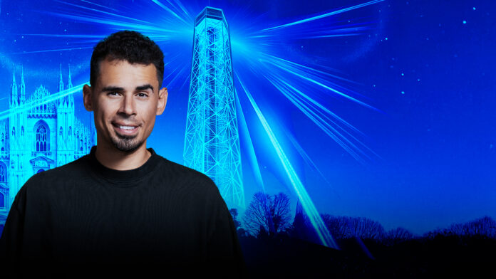 Milano capitale della nightlife al JustMe arriva Afrojack - nella foto l'artista con capelli corti e barba incolta sorride, indossa una maglia nera, sfondo blu con il duomo di MIlano disegnato dietro e un faro che fa luce