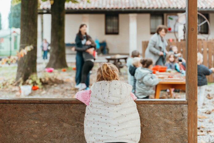 Scuola di infanzia - una nimba bionda con giubbotto bianco con cappuccio è di schiena appoggiata a un muretto e guarda altri bambini giocare in un parco