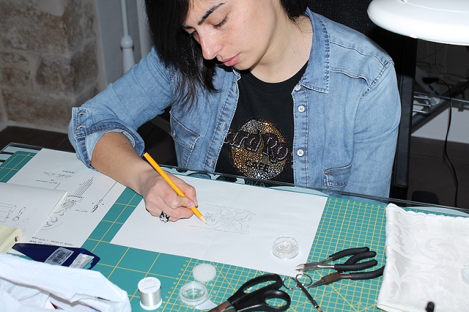 Il tocco di Minerva - una donna con giacchetta di jeans è seduta a unt avolo e sta disegnando un modellino di moda
