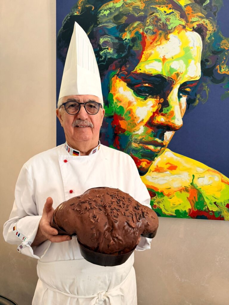 Colombe artigianali - un uomo con baffi e occhiali indossa un cappello bianco dia chef, camicia da chef e tiene in mano un dolce di pasqua 