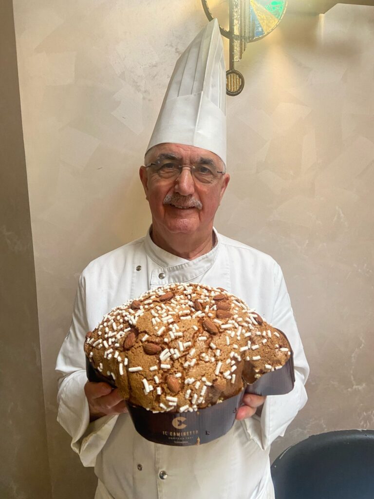 Colombe artigianali - un uomo con baffi e occhiali indossa un cappello bianco dia chef, camicia da chef e tiene in mano un dolce di pasqua