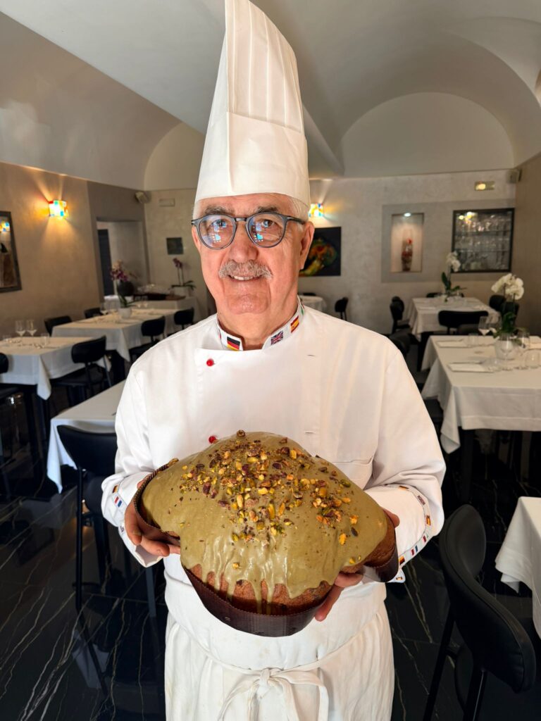 Colombe artigianali - un uomo con baffi e occhiali indossa un cappello bianco dia chef, camicia da chef e tiene in mano un dolce di pasqua 