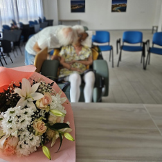 il cancro e le sue storie di cuore - un mazzo di fiori incartato in carta rosa, in primo piano, sfocato sullo sfondo una donna con calze bianche in carrozzina e un uomoche si china e la bacia, in una stanza con delle sedie azzurre