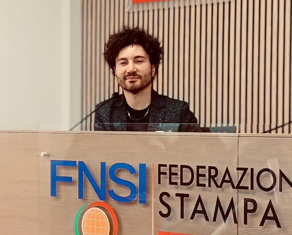 LAZIOSound Gianni Testa con capelli folti ricci neri, barba e pizzetto, è seduto dietro un tavolo conferenza della fnsi