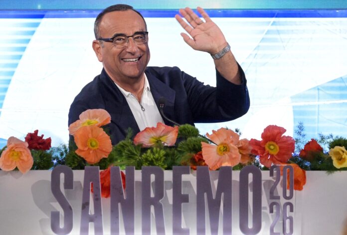 Sanremo TOP seconda serata - Carlo Conti molto abbronzato saluta sorridente, indossa occhiali da vista, camicia bianca aperta sul collo e giacca nera, è seduto e davanti a lui ci sono dei fiori e la scritta Sanremo 2026