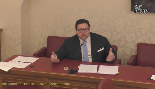 Massimiliano Albanese indossa giacca e cravatta camicia bianca, è seduto ad una scrivania e sta parlando