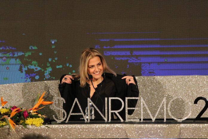 Canzonissima quarta puntata - nella foto Irene Grandi, bionda si sta togliendo una giacca nera, è seduta dietro un banco conferenza e davanti aha la scritta Sanemo