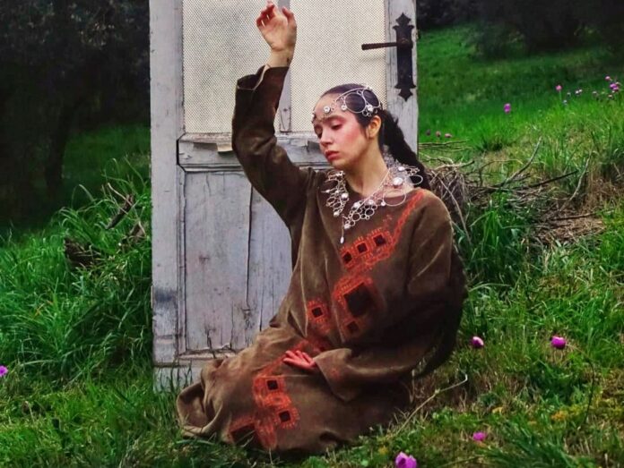 Onirico, il nuovo singolo di Angelica Del Nibbio - Zetatielle Magazine onirico - angelica del nibbio indossa una tunica marrone ed è fotografata all'aperto su un prato, davanti a una porta di legno bianco