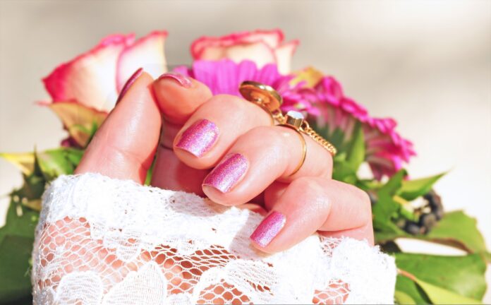 Smalto primavera 2026 la manicure diventa linguaggio di stile - delle rose sullo sfondo e in primo piano una mano di donna con unghie rosa, la mano è in un guanto bianco ma le dita sono scoperte