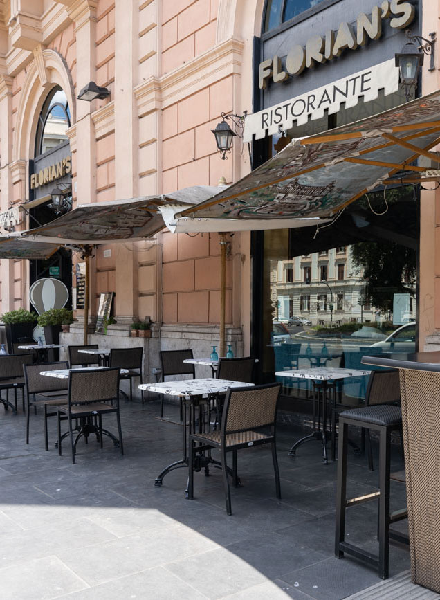 dei tavolini in fila sul marciapiede attaccati al muro di un bar