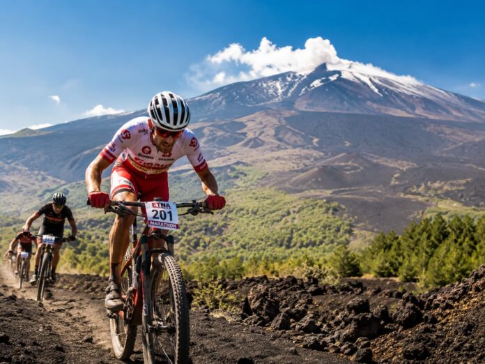Etna Marathon 2026 rinviata a settembre ecco cosa è successo - zewtatielle magazine - immagine generata con IA Etna Marathon, un ciclista su mountain bike su un sentiero di montagna aperto e sullo sfondo l'Etna con delle nuvole che coprono la cima del vulcano. Il biker indossa casco protettivo e tuta da ciclista, sulla bici il cartellino bianco con il numero 201 e la scritta