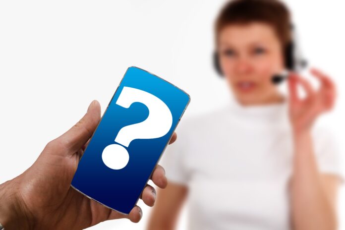 Frodi telefoniche - in primo piano una mano tiene un cellulare aperto su una schermata con un punto interogativo blu e sullo sfondo sfocato si percepisce la sagoma di una donna che indossa cuffia microfono di un call center