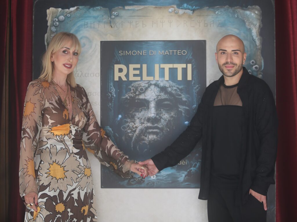 Relitti - nella foto una donna sulla sinistra, con capelli biondi tiene la mano ad un uomo sulla destra e in mezzo a loro il cartellone della mostra
