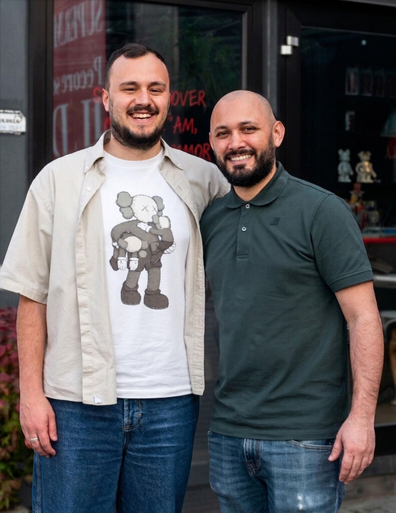 progetto pizza terapia: due uomini, quello a destra calvo con barba, t shirt verde e sorride, quello a sinistra molto alto e corpulento, con capelli neri coriti, barba, indossa jeans e maglietta con sopra una camicia aperta e sorride