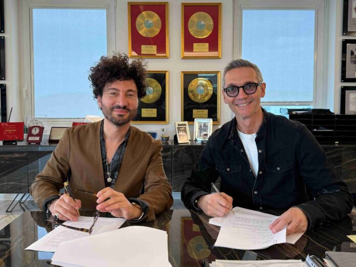 The Saifam Group Gianni Testa è il nuovo Arts Strategic Project Manager - zetatielle Magazine The Saifam Group Gianni Testa - nella foto a destra Gianni Testa con capelli ricci folti sorride e a sinistra Simone Farina con capelli corti e occhiali da vista sorride. Entrambi sono seduti a una scrivania con tanti fogli, uno sta firmando uno foglio l'altro hga occhiali in mano, dietro di loro dei quadri con dei dischi d'oro