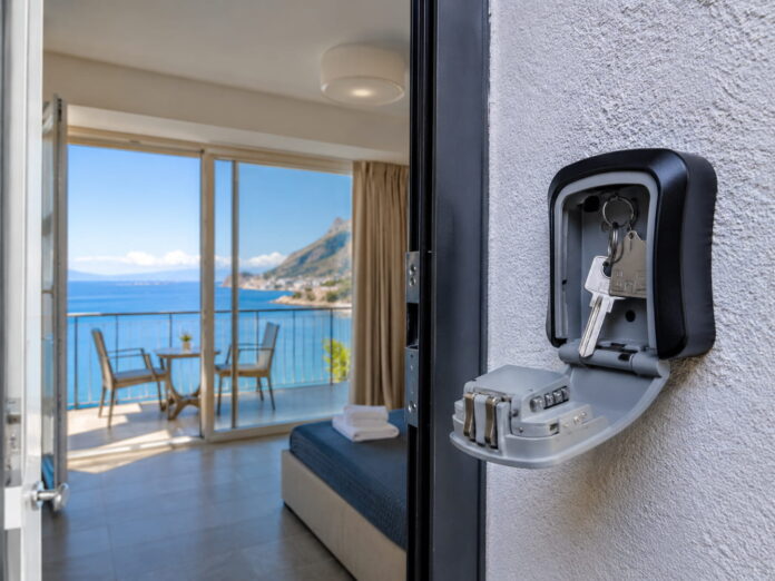 Case vacanze affitti brevi - in primo piano sulla destra una scatola nera con il coperchio aperti e dentro la scatola un mazzo di chiavi, la keybox è davanti all'ingresso di un appartamento con la porta aperta che fa vedere l'interno con una grande vetrata da cui si vede il balcone con tavolino e due sedie, che affaccia sul mare - immagine generata con IA