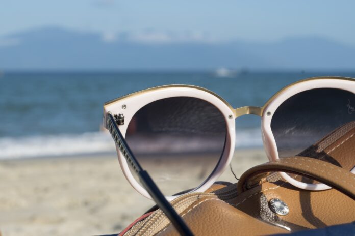 Occhi secchi e il caldo cosa fare e come prevenire - in primo piano degli occhiali da sole appoggiati sul bordo di una borsetta e sullo sfondo spiaggia e mare