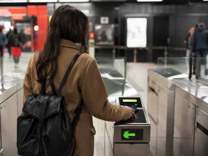 Truffa della metro a Milano e Roma, attenzione al tap out - zetatielle nagazine - immagine genrata con IA truffa della metro - una donna indossa cappotto e zaino sulle spalle. Ha capelli neri lunghi, è di schiena e sta passando la carta sul dispositivo di entrata ai tornelli della metro. Davanti a lei ci sono altre persone - immagine generata con IA