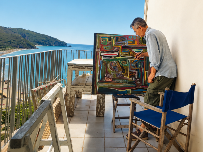 Arte a Sperlonga Fernando Fragione inaugura la mostra Anamnesis - zetatielle magazine - Foto 1 Arte a Sperlonga - nella foto un balcone grande con muri bianchi e ringhiera blu, sul balcone dei cavalletti da pittore e su uno è posato un quadro astratto e un uomo sta dipingendo, dal balcone si vede un bellissimo panorama di mare e costa