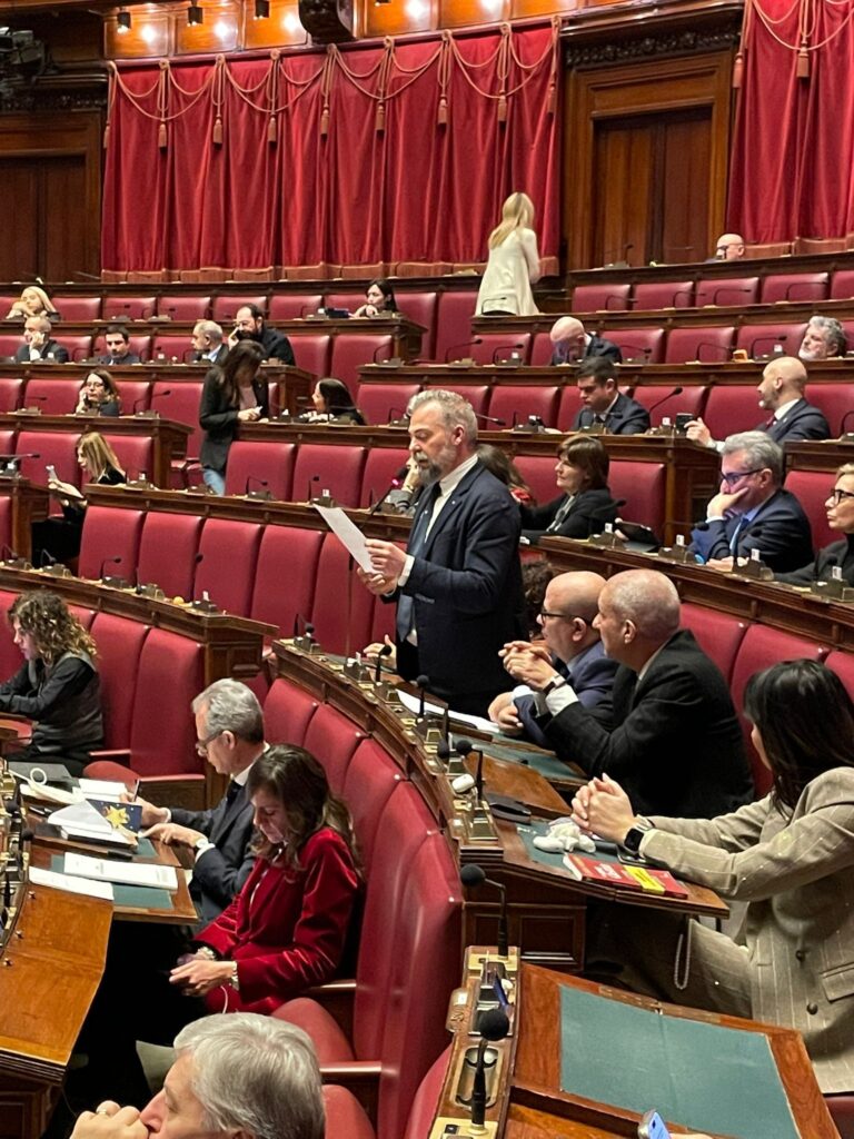 Emiciclo della Camera dei Deputati con poltrone e tende bordeaux e, tra diverse persone sedute, un uomo in piedi in giacca e cravatta legge un discorso.