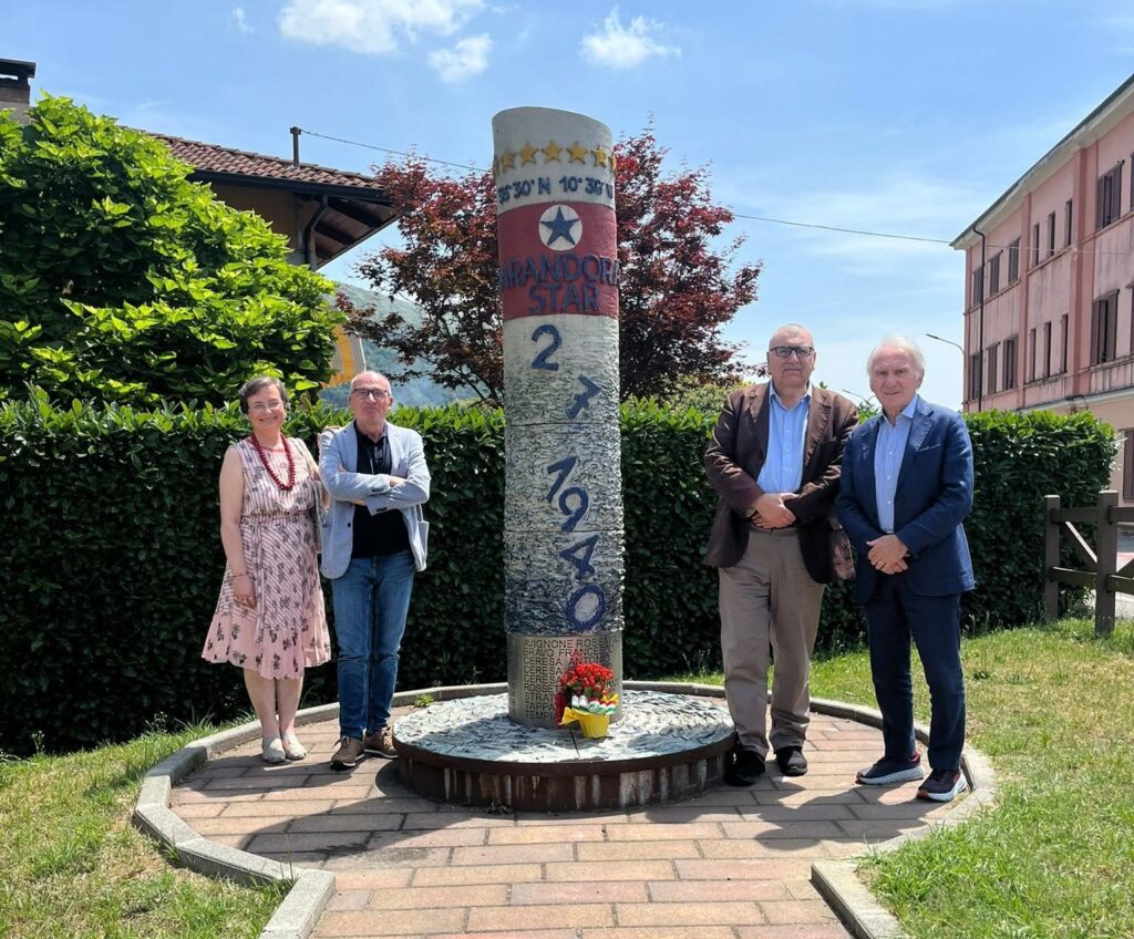 Foto di monumento con fumaiolo dell'Arandora Star, in un'area verde, e quattro persone accanto, due a destra e due a sinistra.