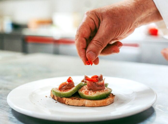 Il nuovo percorso degustazione dello chef Nazareno Fontana, solo a Terracina - zetatielle magazineContrasti d’estate 1 Il nuovo percorso degustazione dello chef Nazareno Fontana, solo a Terracina - nella foto su un piatto bianco delle tartine con delle fette di fico con carne e peperoni e una mano sta aggiungendo dei pezzettini di verdura rossa
