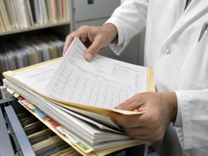 la cartella clinica - nella foto si vede una pila di fascicoli e carte e due mani tengono in mano del fogli, appartengono a una persona in camice bianco - immagine generata con AI