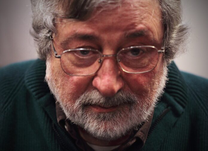 Guccini De Andrè - Francesco Guccini con il volto in primo pian, ha barba incolta, indossa occhiali da vista e ha un'espressione perplessa