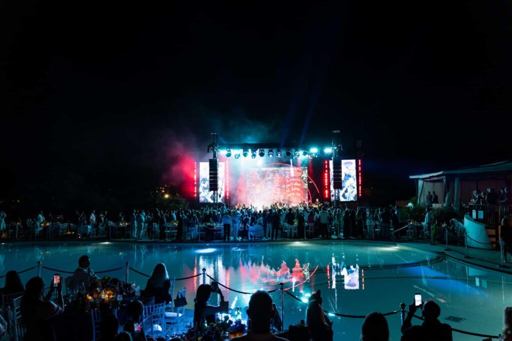 Il BIG Art Festival conquista Porto Cervo con Robbie Williams - un palco illuminato da luci colorate, davanti l palco tanta gente, dietro la folla una piscina. Scena di notte