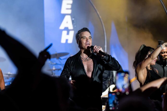 Il BIG Art Festival conquista Porto Cervo con Robbie Williams - zetatielle magazine (2) Il BIG Art Festival conquista Porto Cervo con Robbie Williams - foto a mezzo busto di Robbie Williams che canta indossando una camicia lucida nera aperta sul petto e tiene il microfono in mano. E' su un palco con tanta gente che balla e alza le braccia